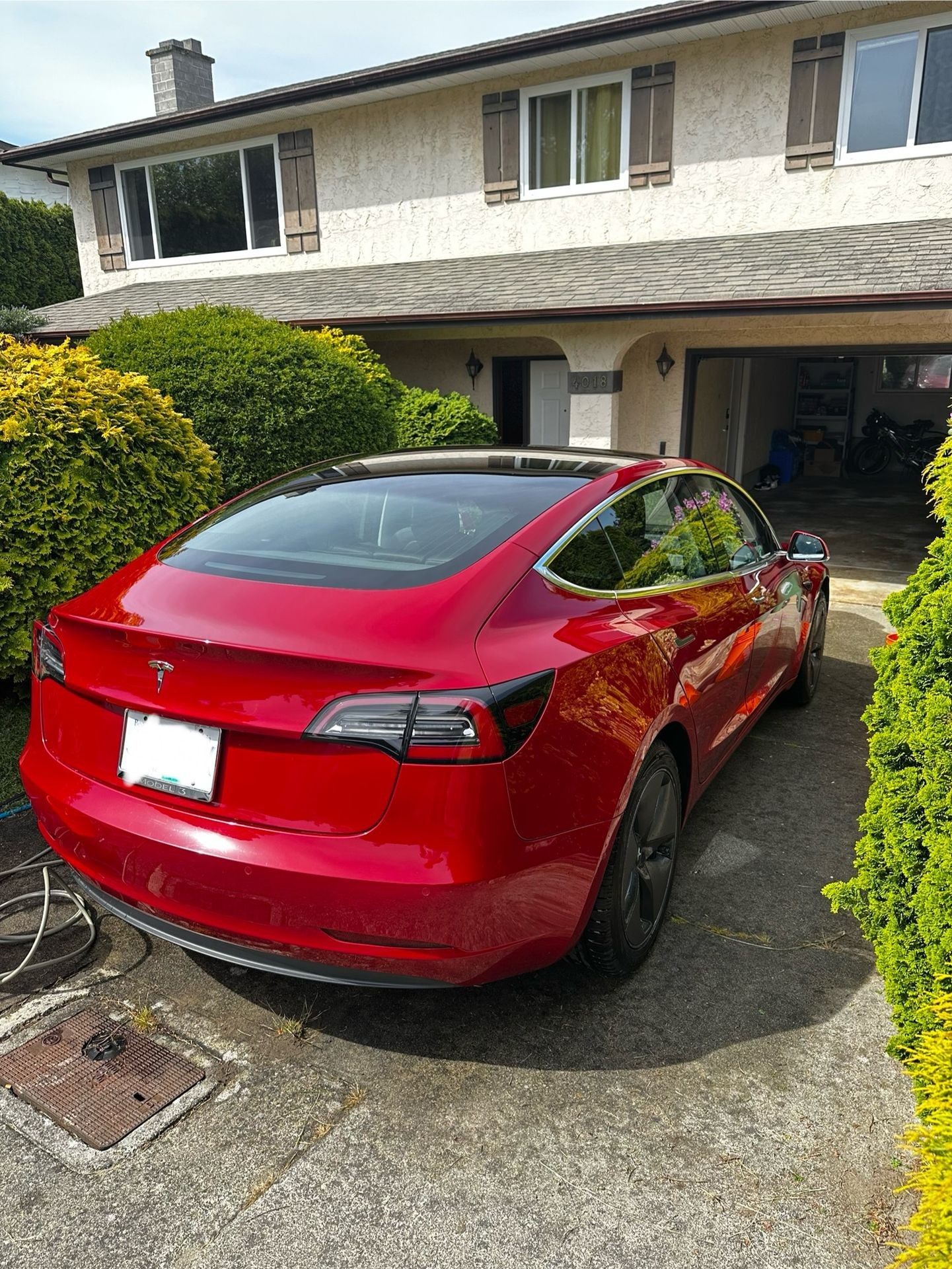 Red Tesla parked in driveway of a suburban house with green shrubs and a garage.